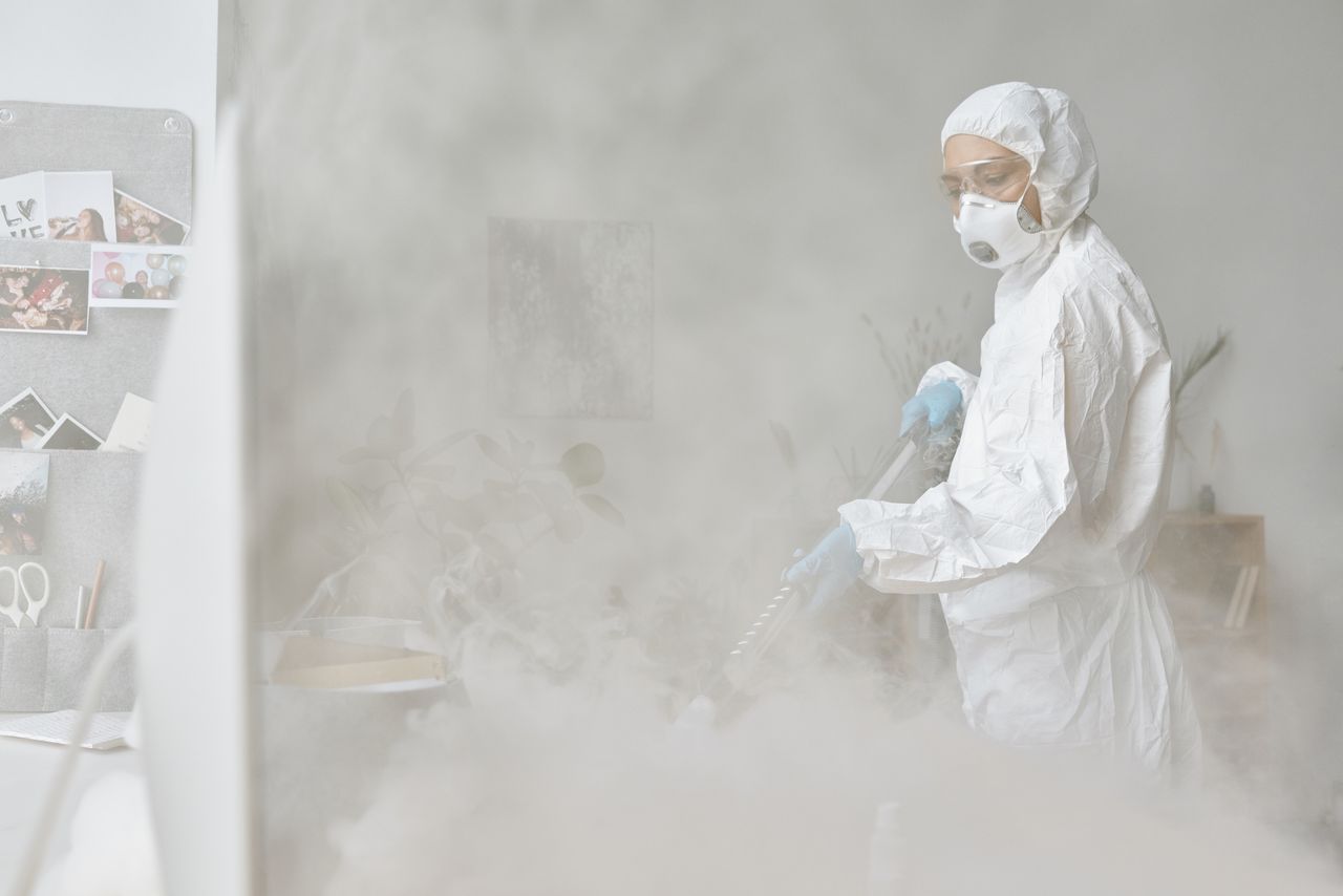 services-04 A woman in a protective suit disinfects an indoor space with fogging equipment.