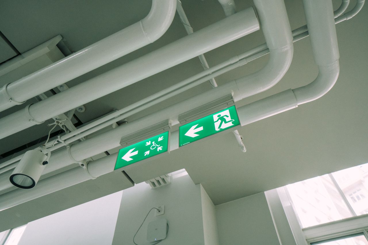 journey White ceiling with industrial pipes and green exit signs in a modern building.