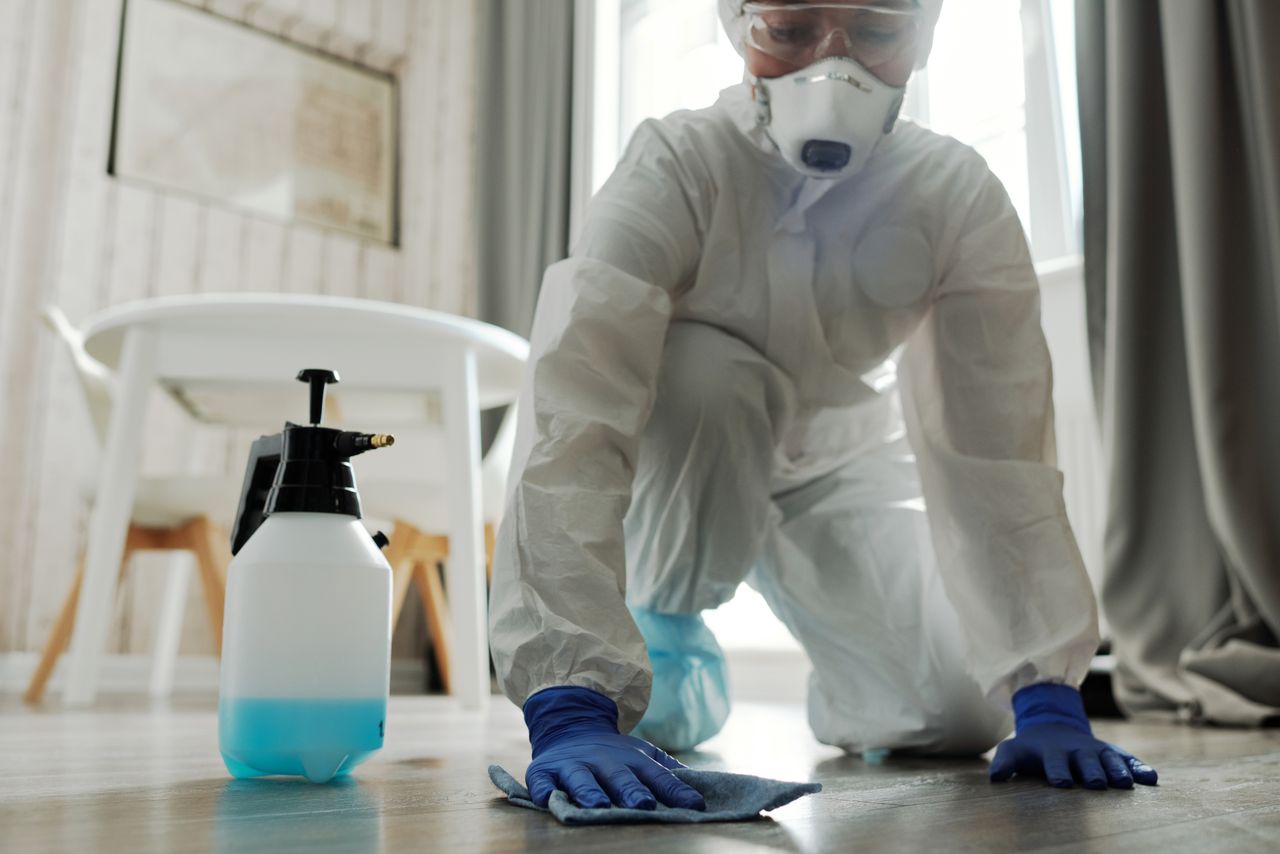 services-01 Person in protective gear cleaning a floor with disinfectant spray and cloth indoors.