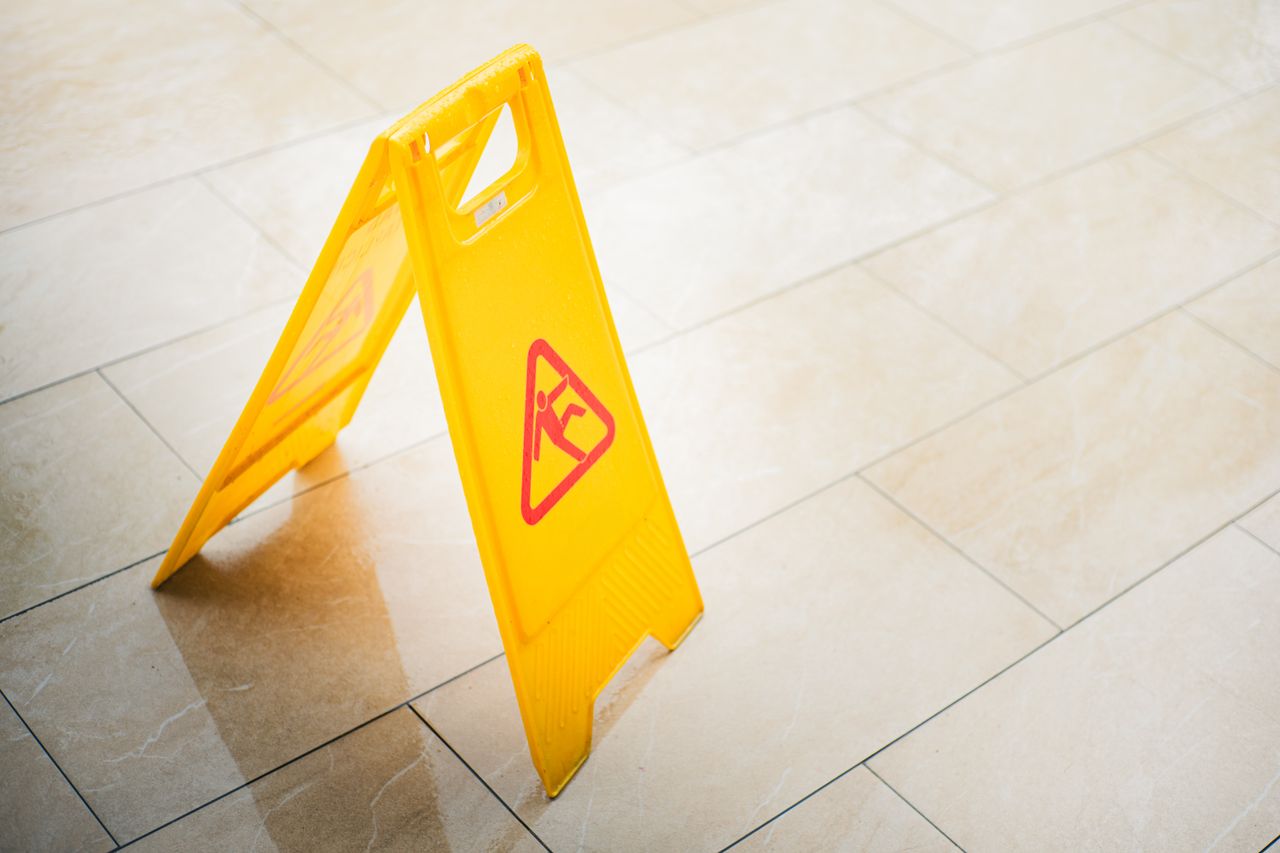 services-03 A bright yellow caution sign on a tiled floor indicates a wet surface, alerting passersby to potential hazards.